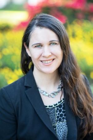 Headshot of Kathreen Thome: a white women with long wavy brunette hair and light colored eyes smiles at the camera. She is wearing a black blazer and a sage green and black patterned shirt, against a backdrop of green bushes with pink flowers.