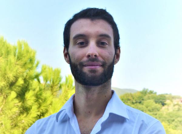 Headshot of Samuele Meschini- a white man with dark hair and a short beard, smiling in front of greenery and a light blue sky. He is wearing a baby blue collared button down.