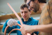 Sam Packman (left) holds a copper VIPER cable. He iw a white man with sandy hair, and is wearing a blue quarter-zip sweater and wearing white nitrile gloves. Nicolo Riva (right) holds he other end of the coil as they inspect it together. He is slightly out of focus, a white man with curly brown hair, glasses, a short beard, and a tan shirt.