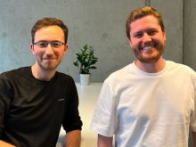 Two scientists looking into the camera and smiling as they sit next to each other at a table.