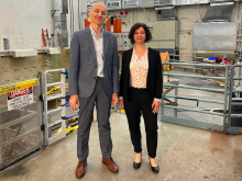 A man and a woman scientist stand next to one another and smile next to one another as they stand in front of a partially-deconstructed fusion device that is housed in a large concrete-lined room on MIT's campus.