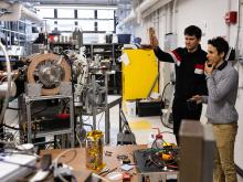 Two men stand on the right side of a sunny industrial lab space pointing to a large piece of research equipment.